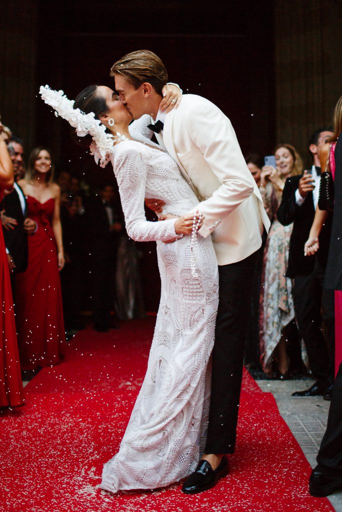 Image depicts a bride and groom embraced in a kiss, standing on red carpet surrounded by their wedding party